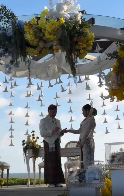 Pasangan mengenakan busana adat Jawa sedang melangsungkan pernikahan di luar ruangan dengan dekorasi bunga kuning dan putih yang indah.