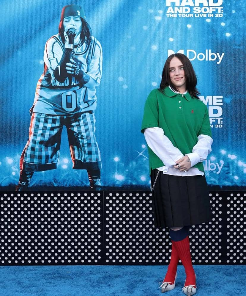 Seorang perempuan berpose di karpet biru di depan poster besar bergambar penyanyi yang sedang tampil di atas panggung.