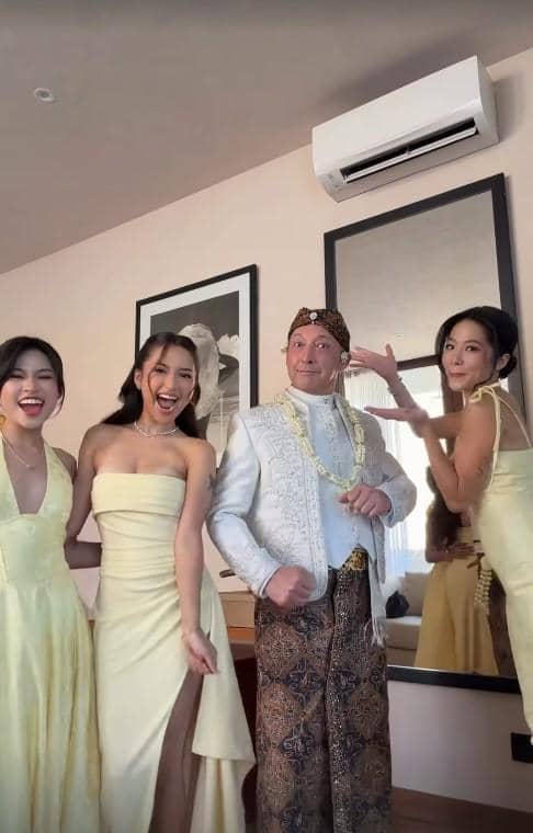 Seorang pria mengenakan busana adat Jawa berdiri bersama tiga wanita bergaun kuning muda yang tersenyum di dalam ruangan.
