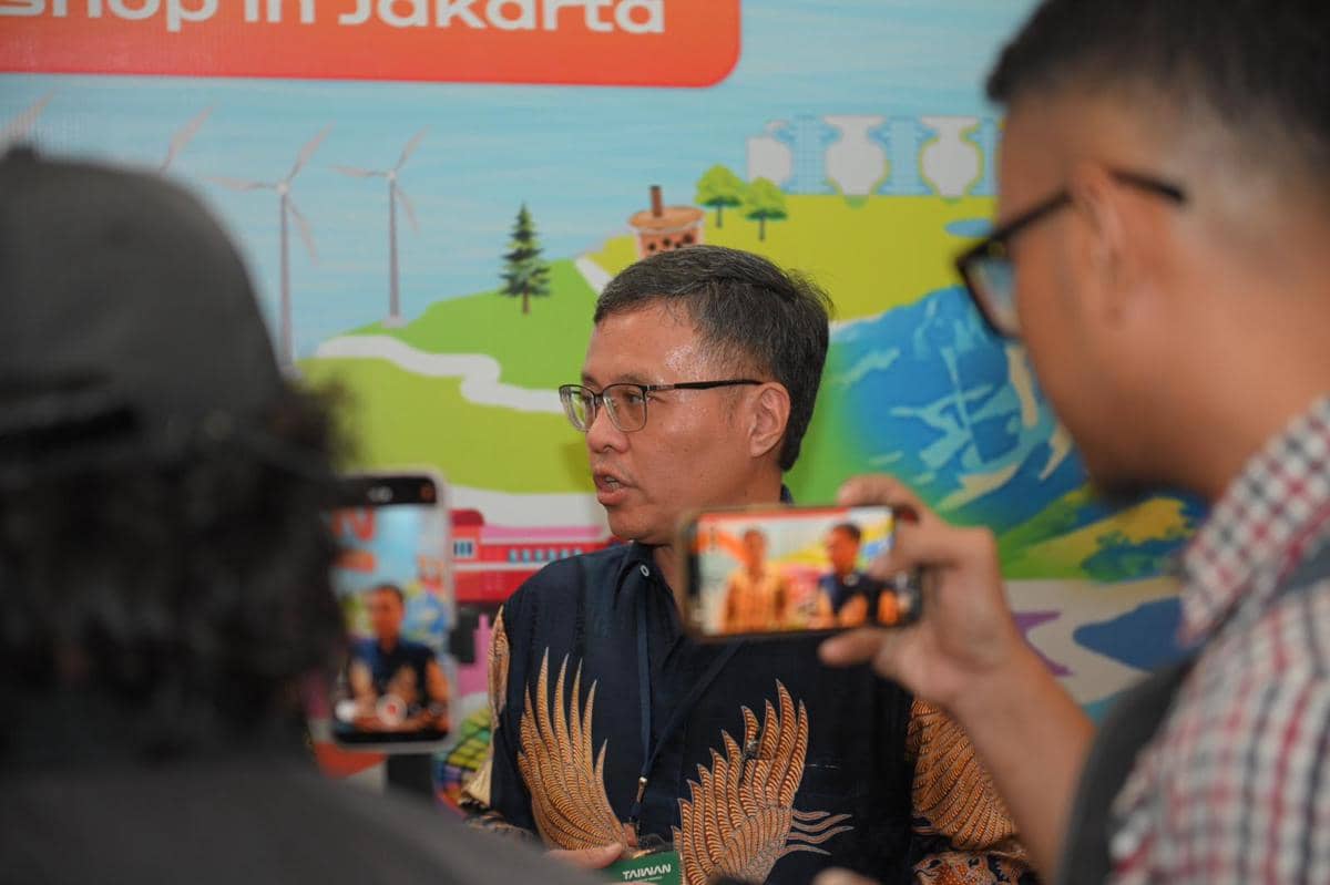 Seorang pria berbicara kepada media di depan latar berwarna cerah bertema pariwisata dengan beberapa orang merekam menggunakan ponsel.