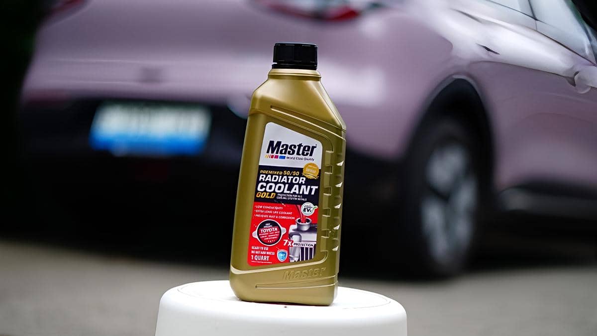 Botol cairan radiator Master Radiator Coolant Gold berwarna emas ditempatkan di atas permukaan putih dengan latar belakang mobil berwarna ungu muda.