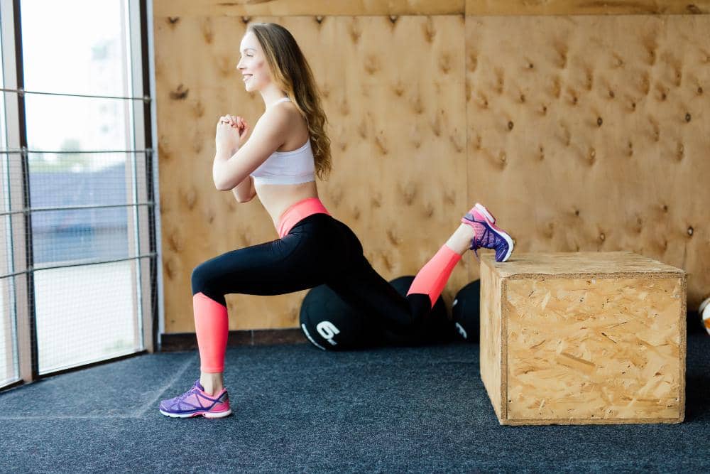 Seorang perempuan berolahraga melakukan gerakan split squat dengan satu kaki di atas kotak kayu di ruang gym dengan pencahayaan alami.