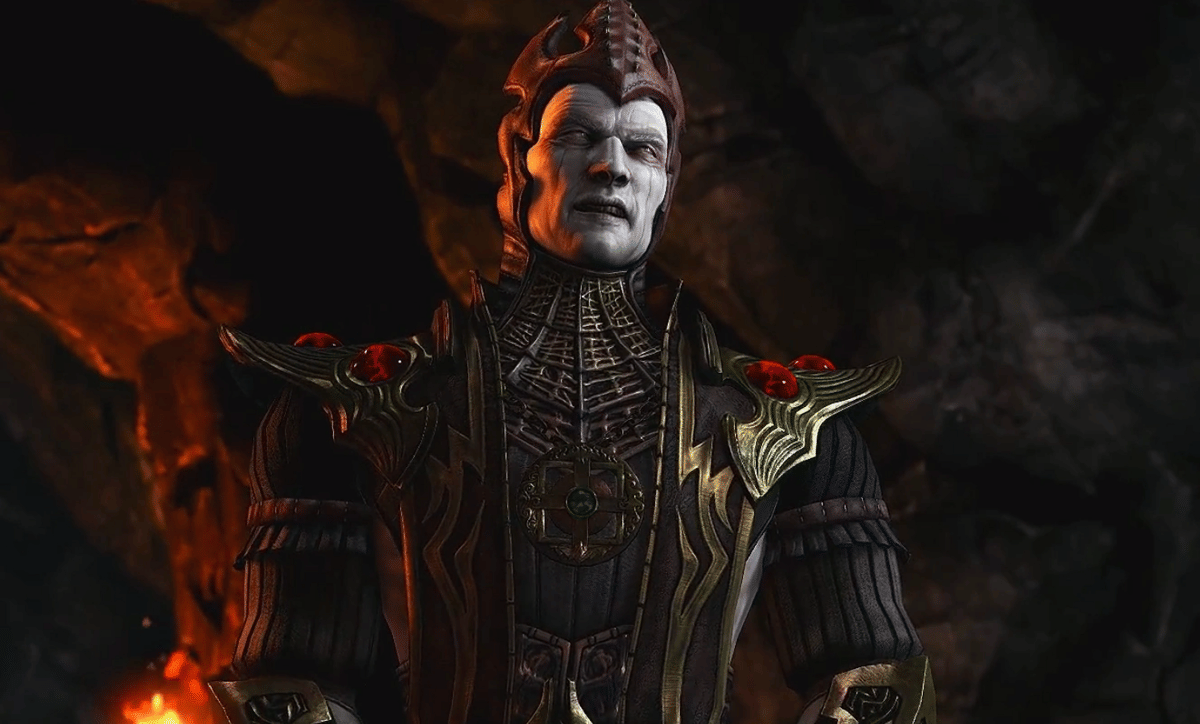Karakter Shinnok dari Mortal Kombat X mengenakan pakaian gelap berhias emas dan batu merah di dalam gua bercahaya api.