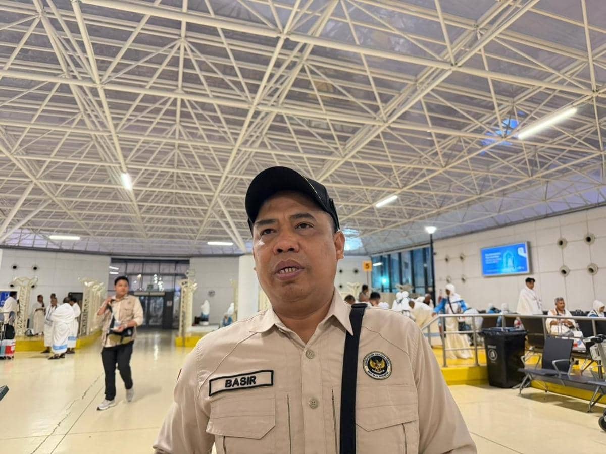 Seorang petugas berpakaian seragam krem dengan topi hitam berada di area bandara Jeddah yang ramai dengan jamaah haji.