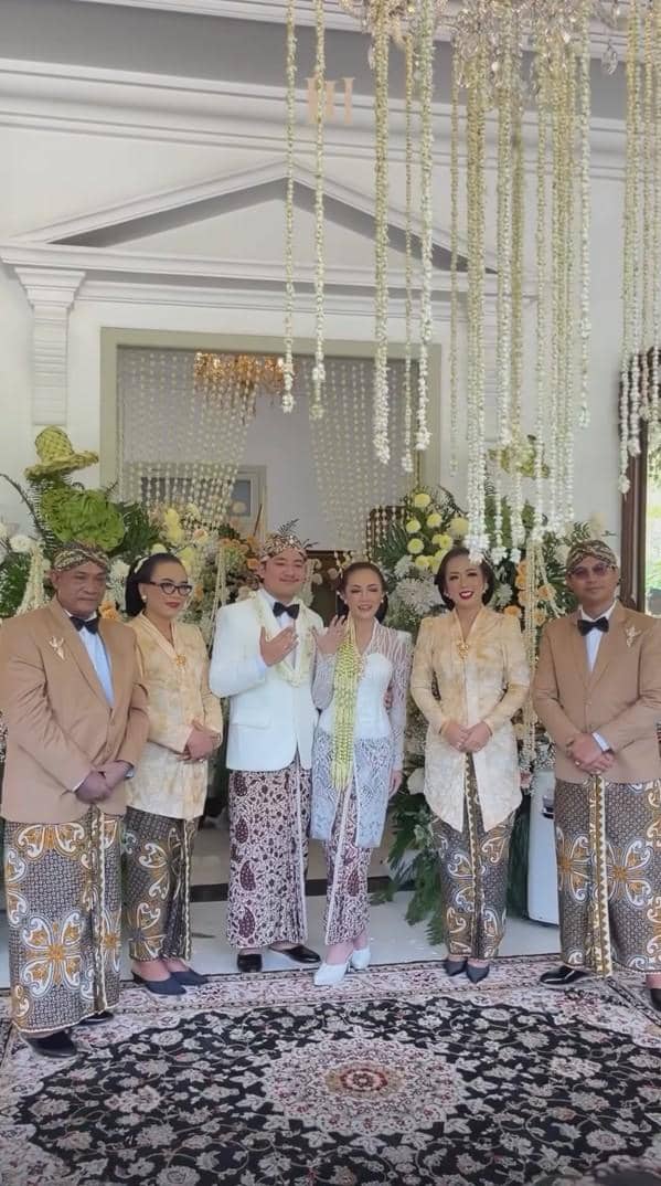 Pasangan pengantin mengenakan busana adat Jawa berfoto bersama keluarga di pelaminan dengan dekorasi bunga dan janur gantung.