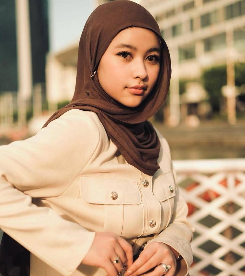 Seorang perempuan berhijab cokelat mengenakan jaket krem berpose di luar ruangan dengan latar belakang gedung modern.