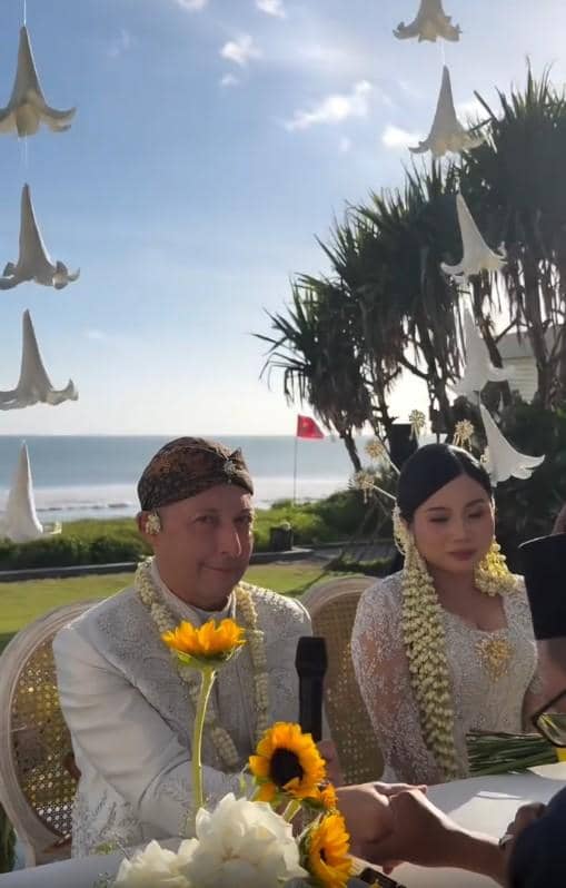 Pasangan pengantin mengenakan busana adat Jawa duduk di pelaminan dengan latar pantai dan dekorasi bunga gantung putih.