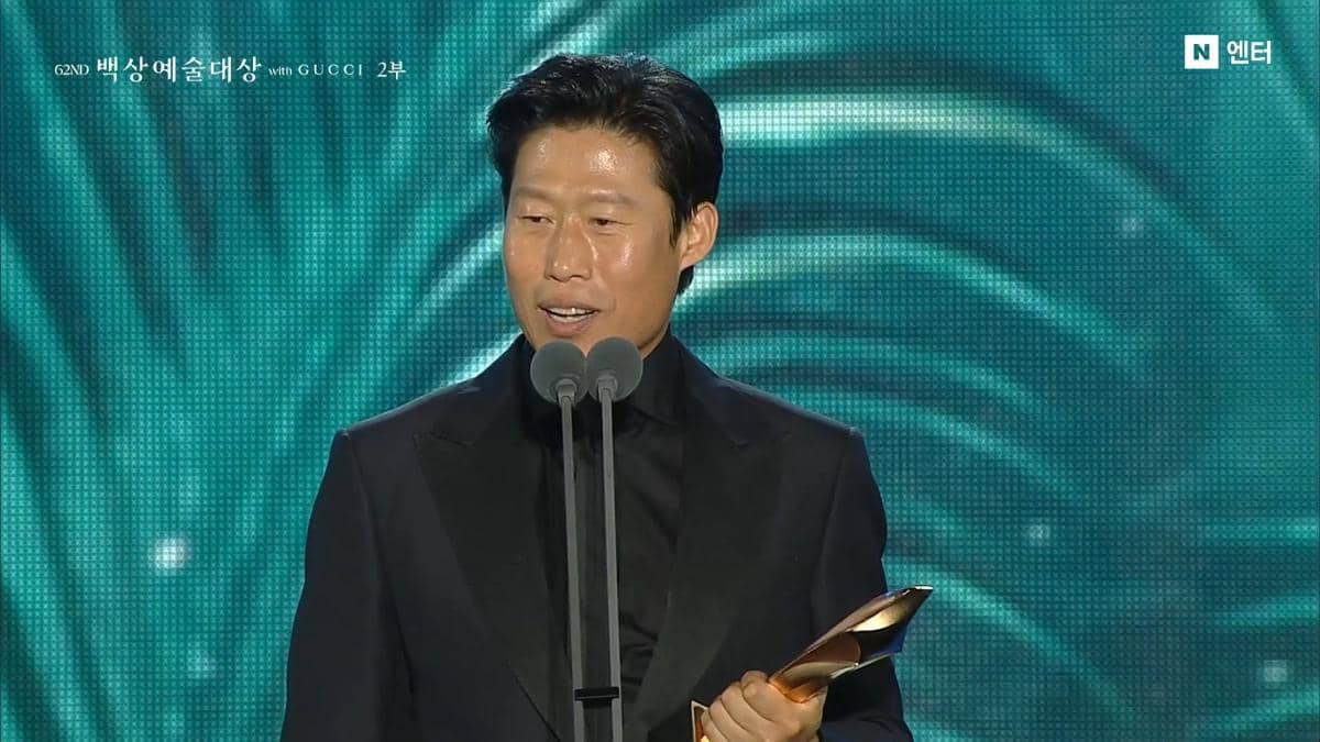 Seorang pria mengenakan setelan jas hitam memegang trofi dan berbicara di depan mikrofon ganda di panggung Baeksang Art Awards 2026.