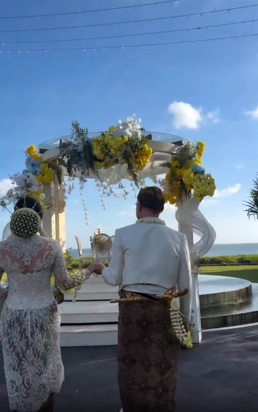 Sepasang pengantin mengenakan busana adat berjalan menuju pelaminan berhias bunga kuning dan putih di tepi pantai saat upacara pernikahan.
