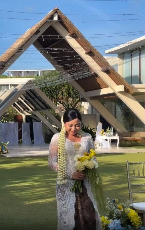 Seorang wanita mengenakan kebaya putih dan kain batik sambil memegang buket bunga kuning di acara pernikahan outdoor dengan dekorasi modern.