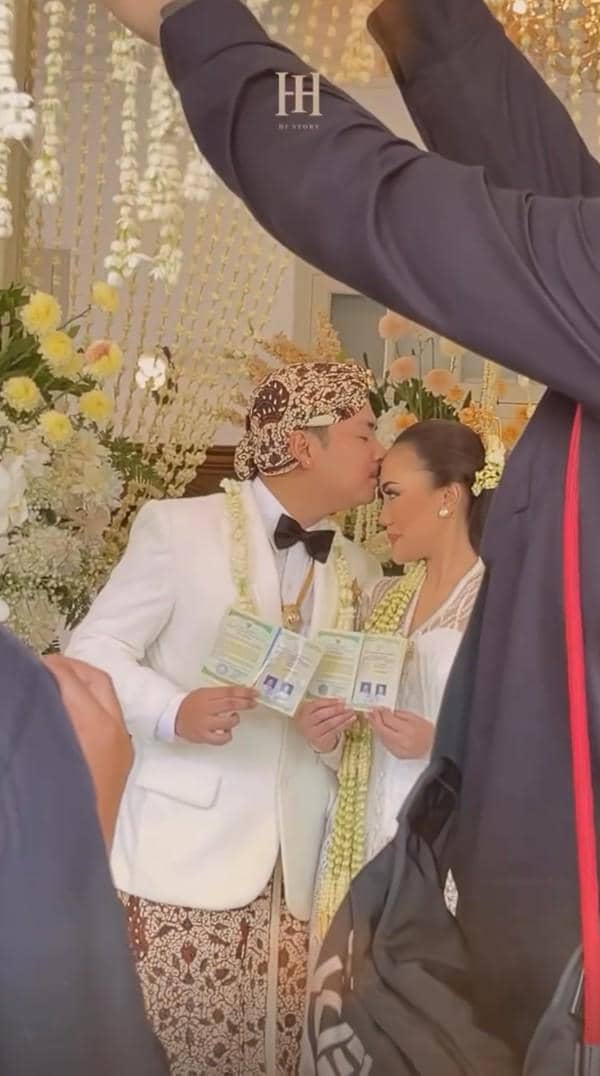 Pasangan pengantin mengenakan busana adat Jawa berwarna putih sambil memegang buku nikah dan berpose di pelaminan berhias bunga.