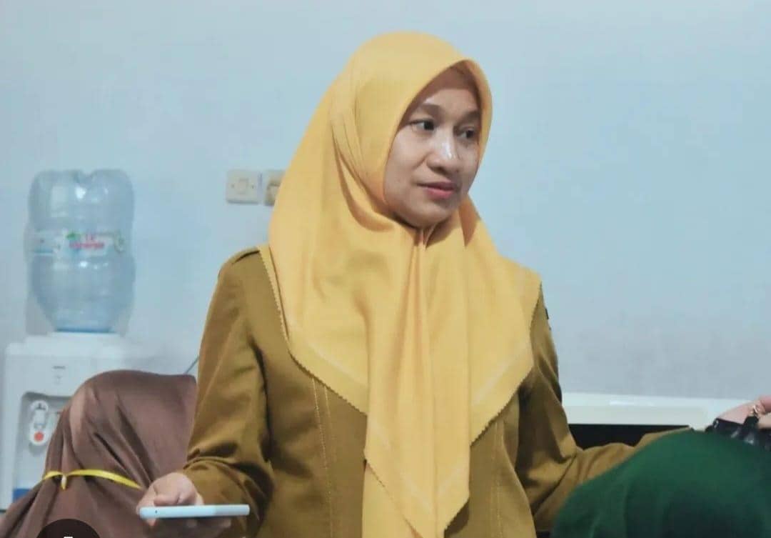 Seorang perempuan berhijab kuning mengenakan seragam cokelat sedang berbicara di dalam ruangan dengan dispenser air di latar belakang.