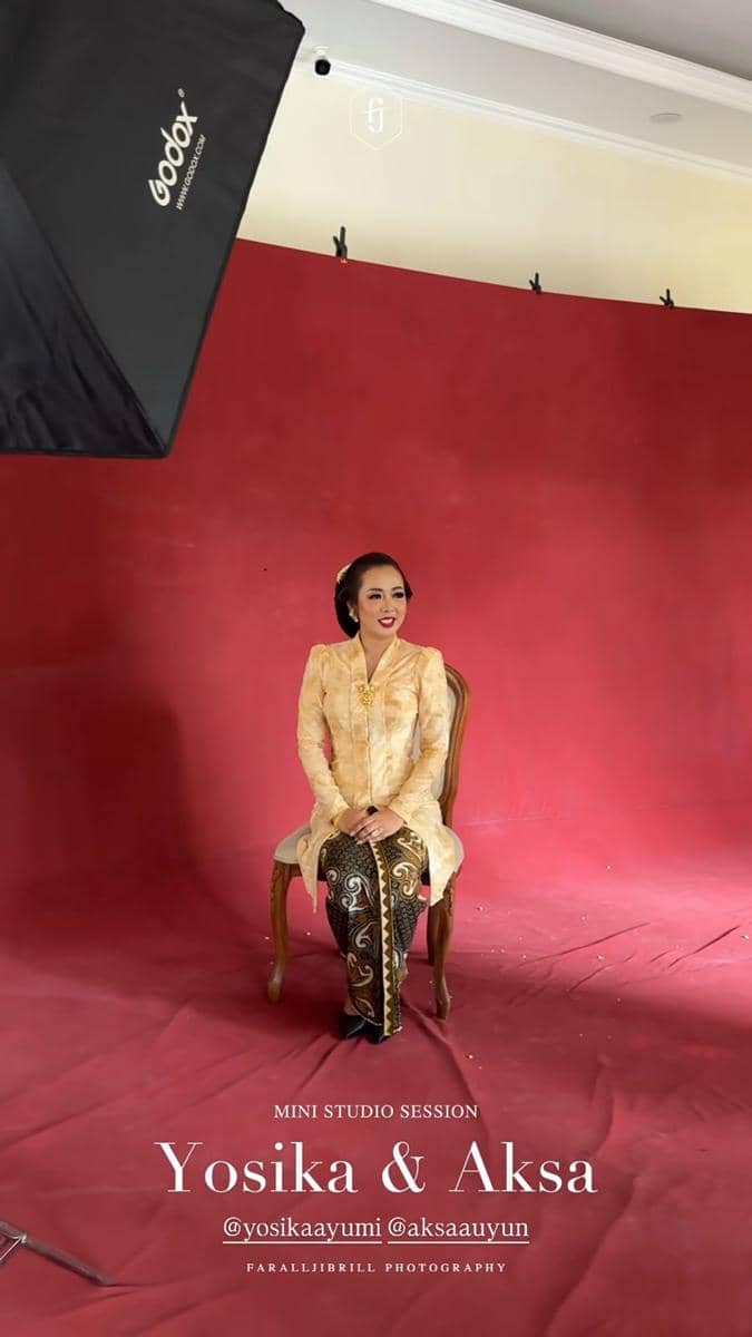 Seorang wanita mengenakan kebaya krem dan kain batik duduk di kursi kayu di studio foto dengan latar merah dan pencahayaan profesional.