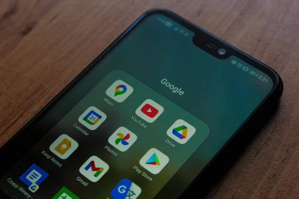 Tampilan layar ponsel pintar menampilkan folder aplikasi Google seperti Maps, YouTube, Drive, dan Gmail di atas meja kayu.