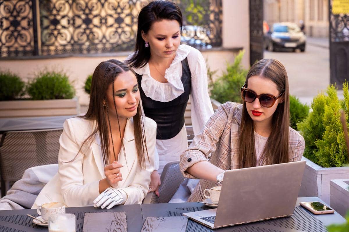 Tiga wanita muda berdiskusi di luar ruangan sambil melihat layar laptop di meja kafe dengan suasana santai dan profesional.