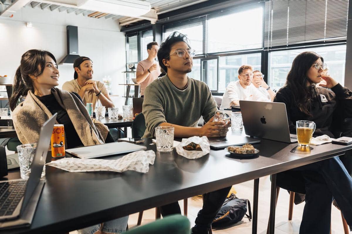 Sekelompok orang duduk di ruang kerja modern dengan laptop dan minuman di meja, tampak fokus mengikuti presentasi atau diskusi.
