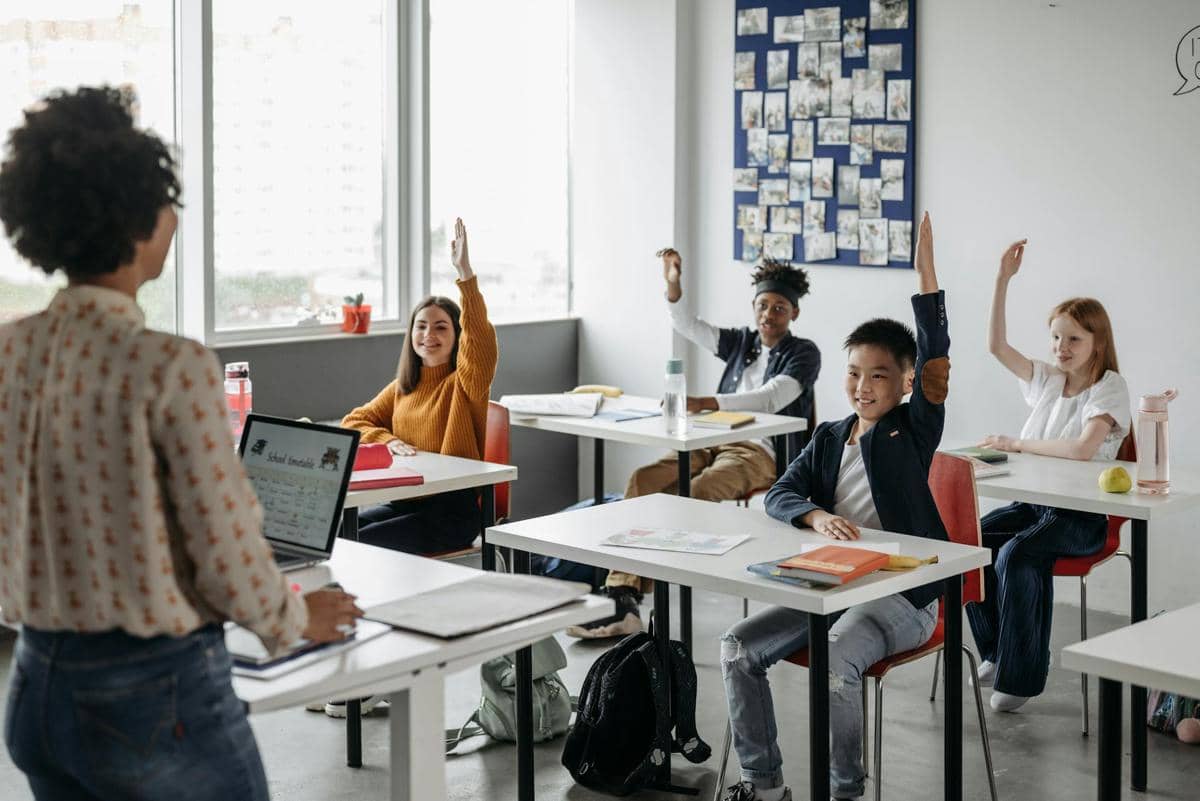 Beberapa siswa duduk di dalam kelas sambil mengangkat tangan untuk menjawab pertanyaan dari guru yang berdiri di depan kelas.