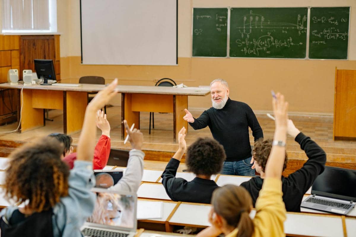 Seorang dosen berinteraksi dengan mahasiswa yang mengangkat tangan di ruang kelas dengan papan tulis berisi rumus di belakangnya.