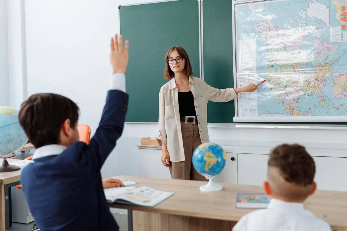 Guru sedang mengajar di kelas sambil menunjuk peta dunia di papan, sementara seorang siswa mengangkat tangan untuk menjawab pertanyaan.