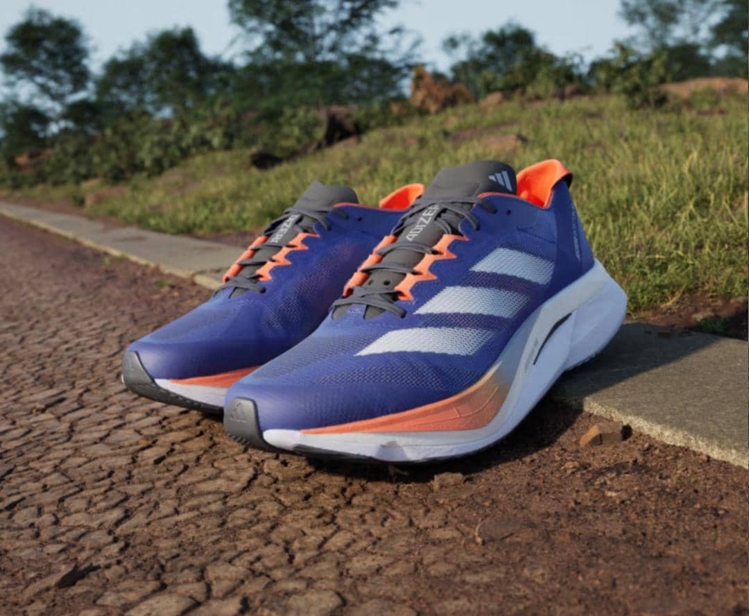 Sepasang sepatu lari Adidas Adizero Boston 12 berwarna biru dengan aksen oranye dan putih diletakkan di jalan beraspal dekat area hijau.