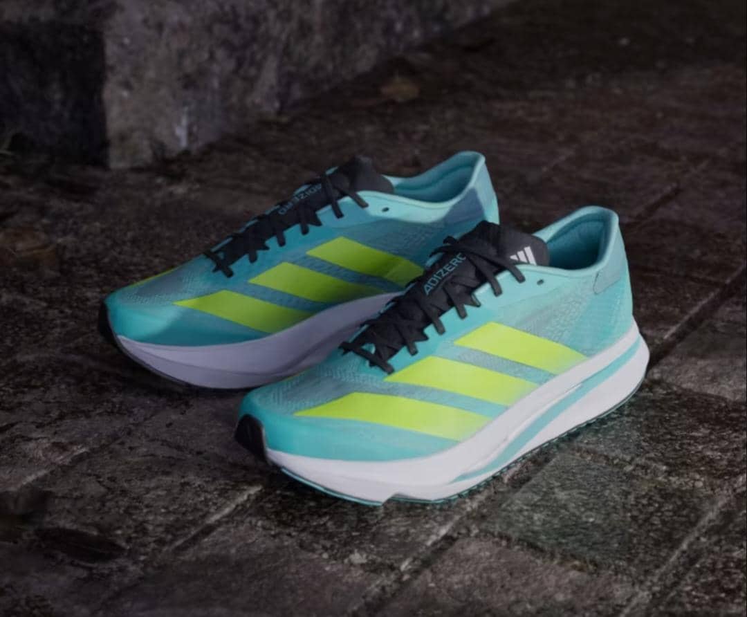 Sepasang sepatu lari Adidas Adizero SL 2 berwarna biru toska dengan garis kuning neon dan sol putih di atas permukaan jalan berbatu.