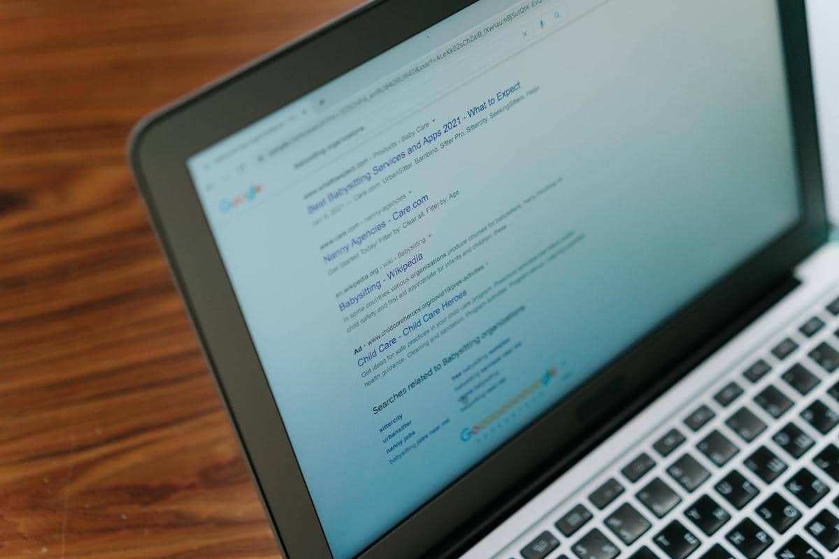 Layar laptop menampilkan hasil pencarian di mesin pencari internet dengan latar meja kayu berwarna cokelat.