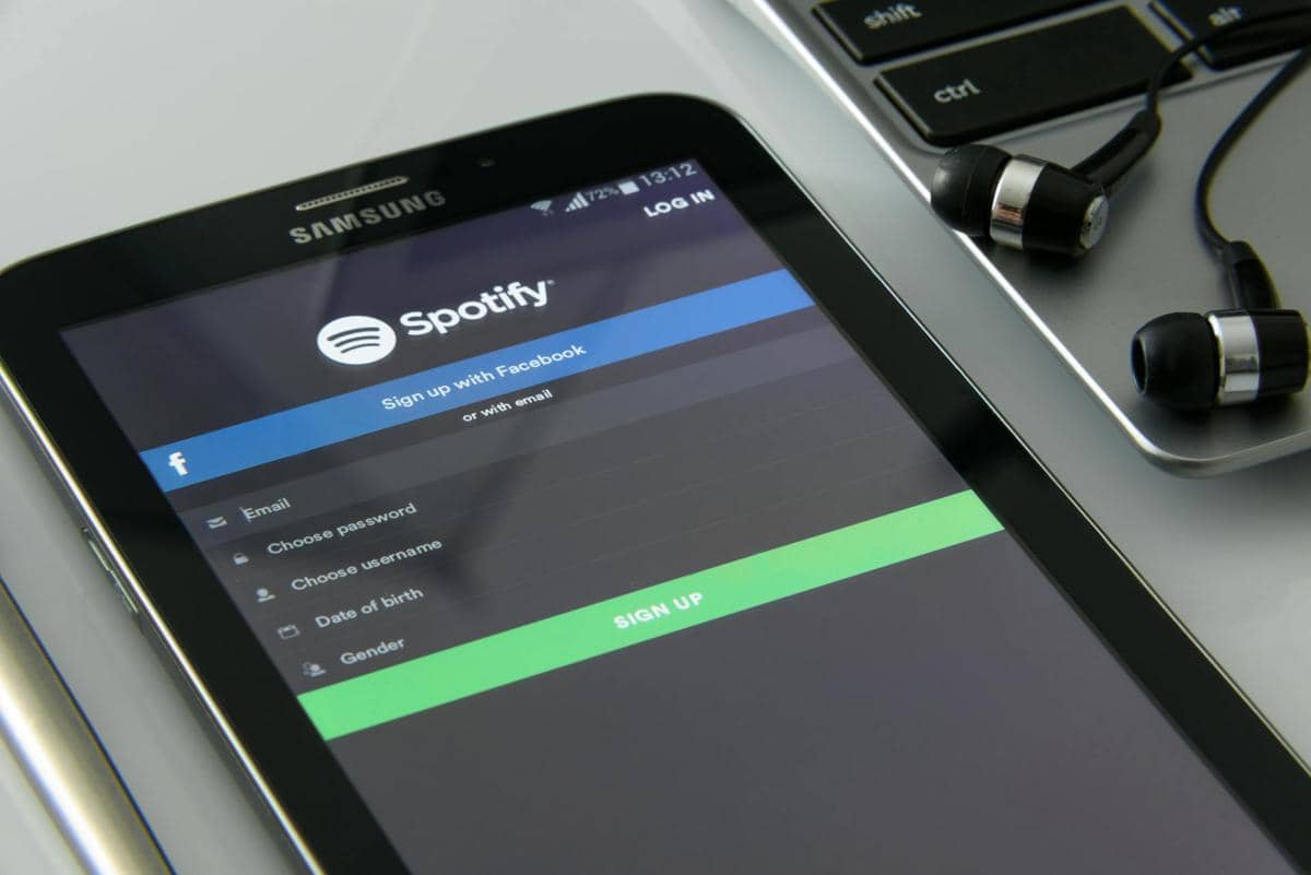 Tampilan layar tablet Samsung menampilkan halaman pendaftaran aplikasi Spotify di samping laptop dan sepasang earphone hitam.