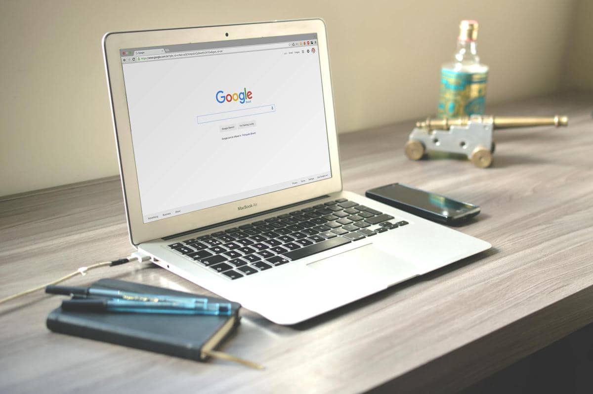 Laptop terbuka menampilkan halaman pencarian Google di atas meja kayu dengan ponsel, buku catatan, dan pena di sekitarnya.