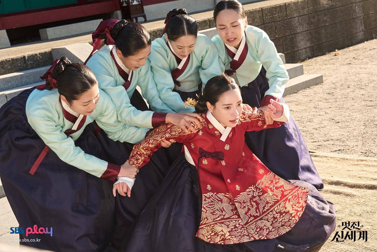 Beberapa wanita mengenakan hanbok tradisional Korea, satu di antaranya berpakaian merah tampak ditahan oleh empat wanita lain di tangga istana.