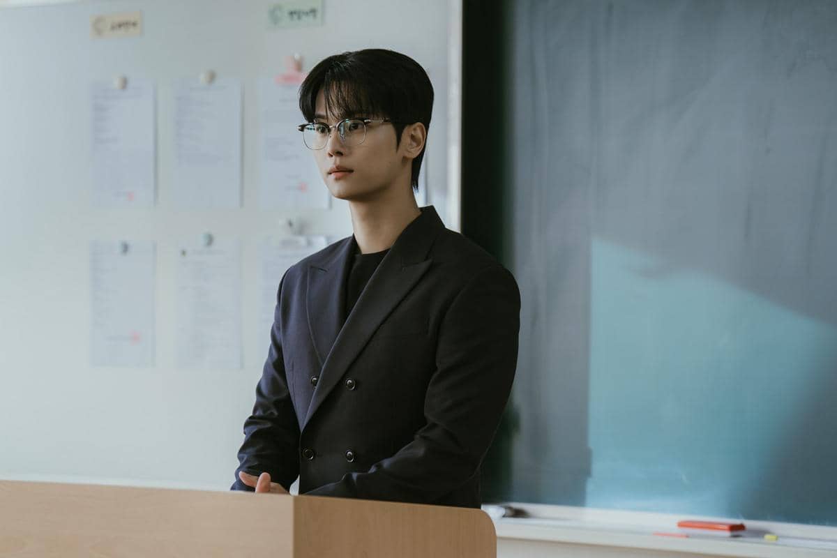 Seorang pria mengenakan setelan jas hitam dan kacamata berdiri di depan podium di dalam ruang kelas dengan papan tulis di belakangnya.