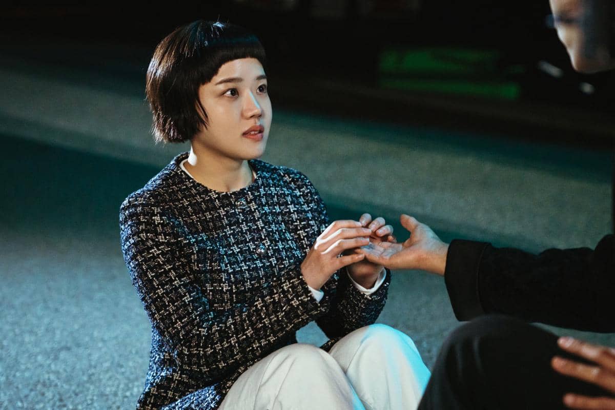 Seorang wanita berambut pendek mengenakan jaket bermotif duduk di jalan sambil memegang tangan seseorang dalam adegan drama Absolute Value of Romance.