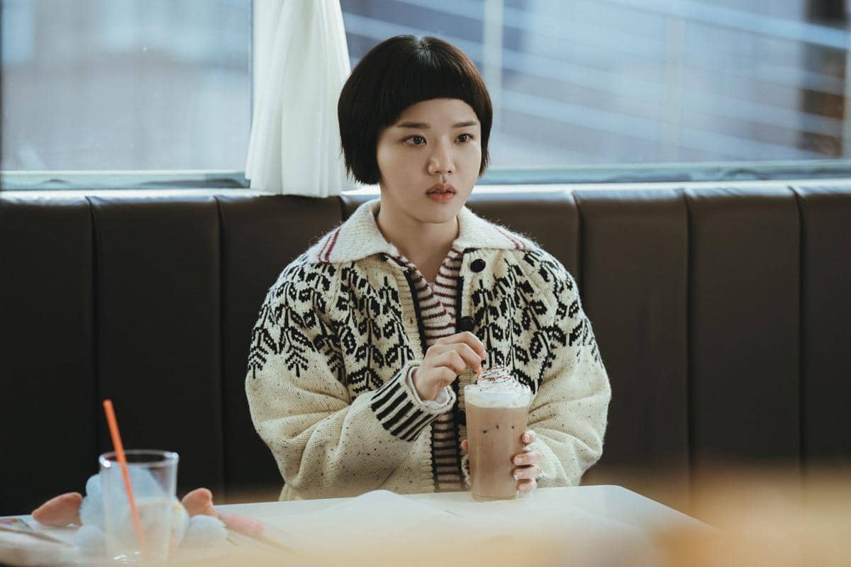 Seorang wanita berambut pendek mengenakan sweter bermotif duduk di kafe sambil memegang minuman cokelat dingin dengan krim di atasnya.