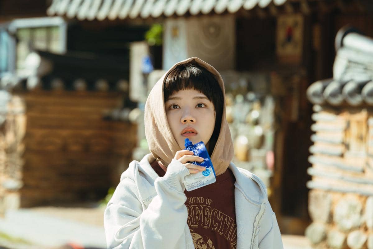 Seorang perempuan muda mengenakan hoodie krem dan jaket abu-abu sedang memegang minuman kemasan biru di luar ruangan dengan latar bangunan tradisional.