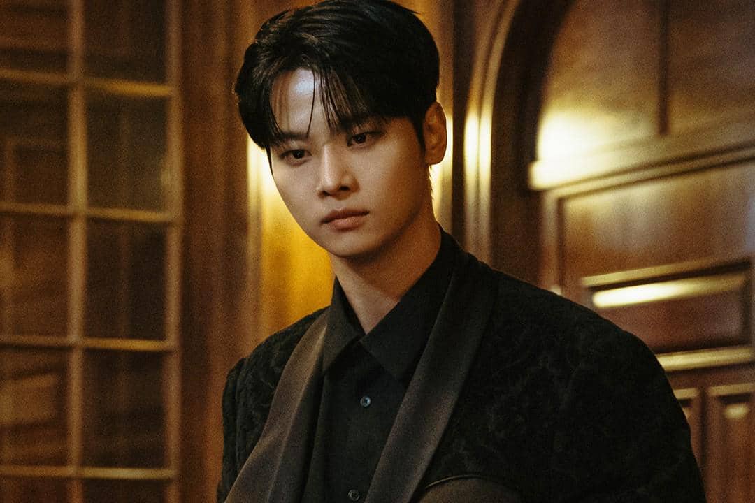Seorang pria berambut hitam mengenakan setelan hitam berdiri di ruangan kayu dengan pencahayaan hangat dalam adegan drama Absolute Value of Romance.