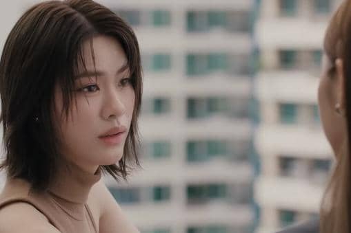 Seorang perempuan berambut pendek menatap seseorang di depannya dalam cuplikan drama Moonshadow dengan latar gedung bertingkat.
