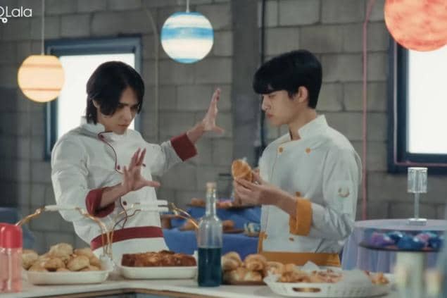 Dua pria mengenakan seragam koki berdiri di dapur roti dengan meja penuh pastry dan roti, salah satunya berpose dramatis di depan yang lain.