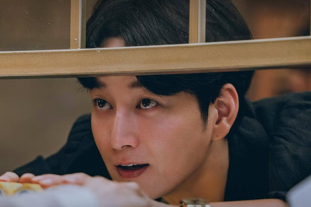 Seorang pria berambut hitam menatap dengan ekspresi lembut melalui jendela kayu dalam adegan drama Gold Land.