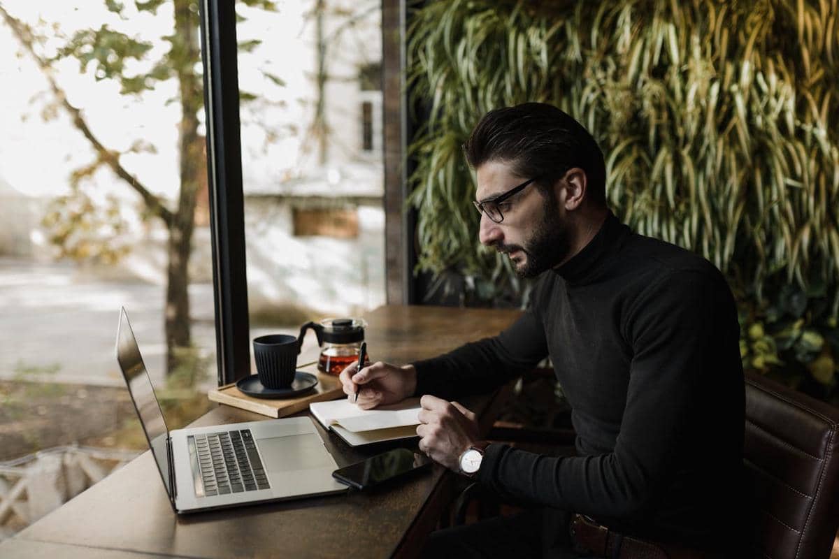 Seorang pria mengenakan sweter hitam bekerja dengan laptop di kafe, mencatat di buku sambil duduk di dekat jendela besar dengan pemandangan luar ruangan.