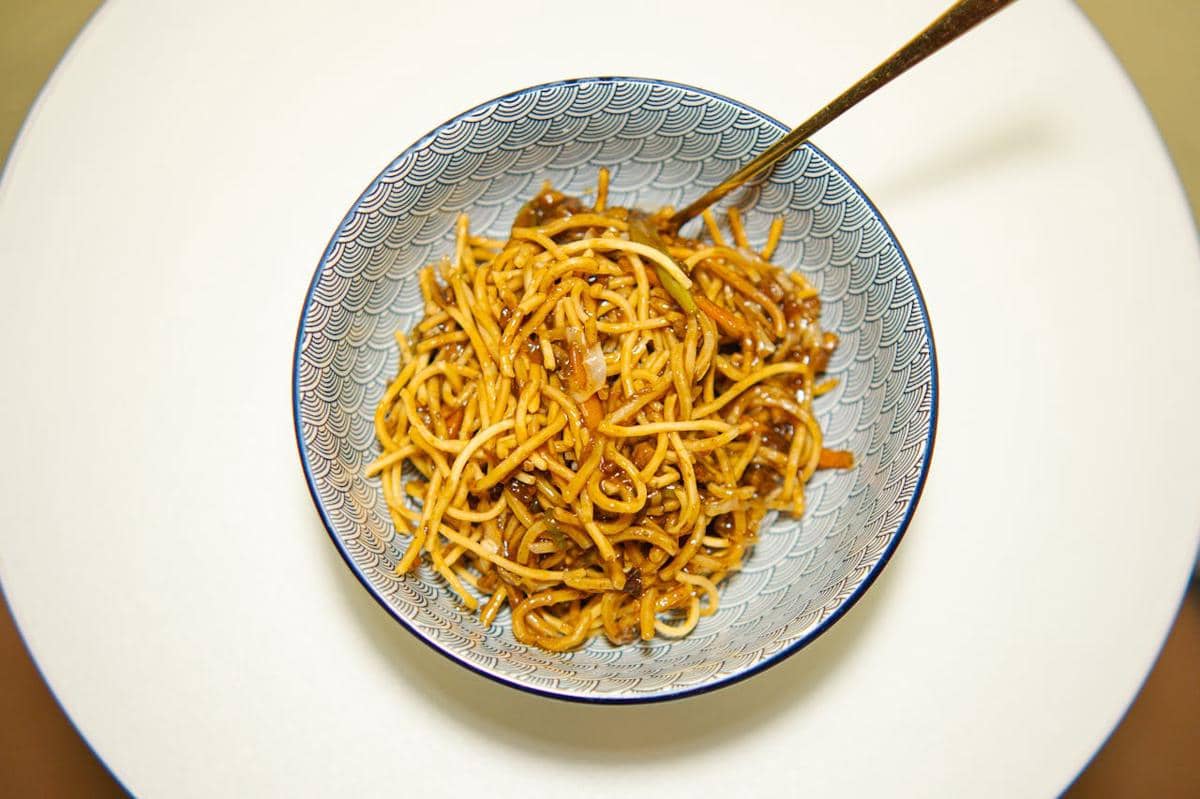 Semangkuk spaghetti dengan saus kecap asin disajikan di mangkuk bermotif biru dengan sendok di atas meja putih.