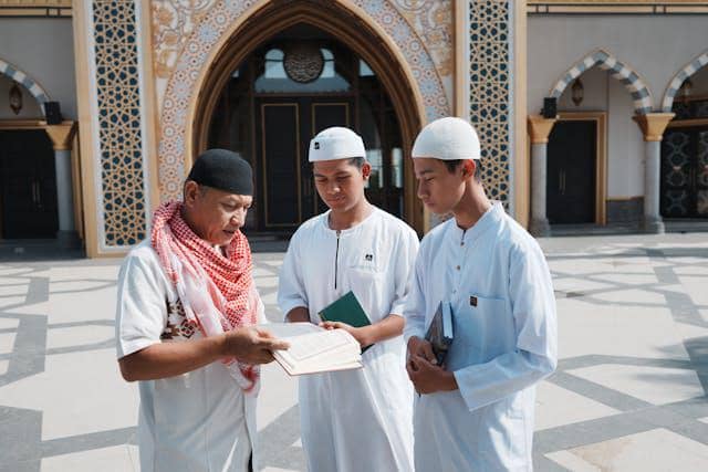 Seorang guru mengenakan sorban merah muda berbicara dengan dua santri berpakaian putih di halaman masjid yang megah.