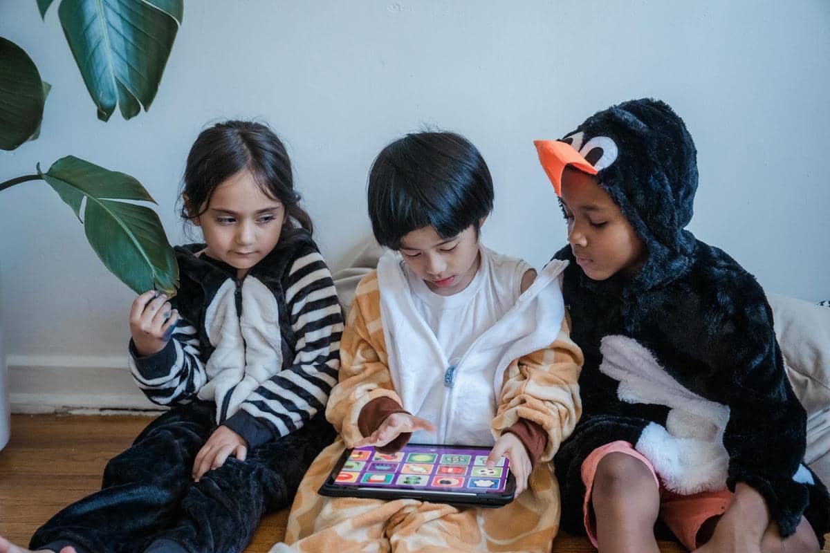 Tiga anak mengenakan kostum hewan duduk bersama di lantai sambil bermain tablet dengan aplikasi berwarna di layar.