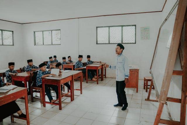 Seorang guru mengajar sekelompok santri laki-laki yang duduk di meja kayu dalam ruang kelas sederhana dengan papan tulis di depan.