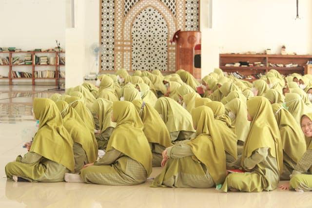 Sekelompok santri perempuan mengenakan seragam hijau duduk bersama di dalam ruangan masjid dengan latar dinding berpola islami.