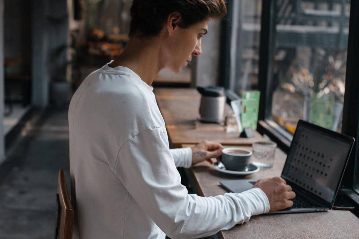 Seorang pria mengenakan kaus putih bekerja dengan laptop di atas meja kayu di kafe sambil memegang secangkir kopi di dekat jendela besar.
