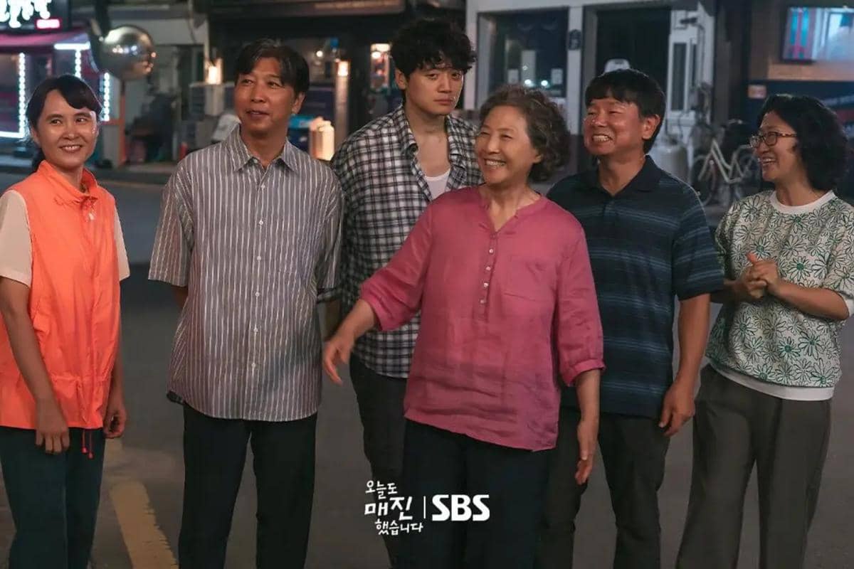 Enam orang berdiri di jalan pada malam hari dengan ekspresi bahagia, salah satunya mengenakan kemeja merah muda, dalam adegan drama Sold Out On You.