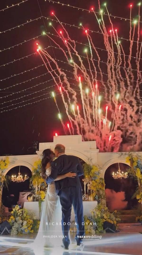 Pasangan pengantin berpelukan di panggung resepsi malam hari dengan kembang api berwarna merah dan hijau di langit belakang mereka.