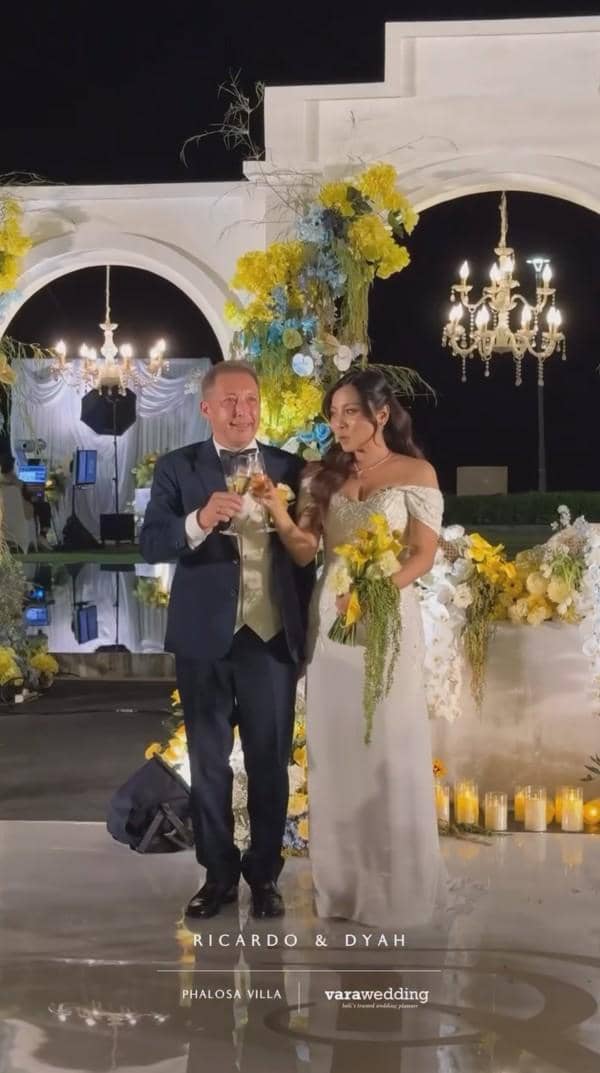 Pasangan pengantin berdiri di depan dekorasi bunga kuning dan putih dengan lampu gantung mewah saat resepsi pernikahan malam hari.