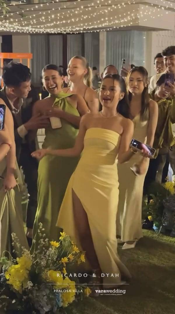 Tamu undangan mengenakan gaun elegan berwarna kuning dan hijau menikmati suasana resepsi pernikahan di bawah lampu hias.