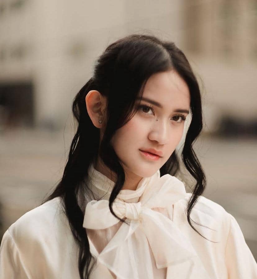 Seorang perempuan berambut hitam bergelombang mengenakan blus putih dengan pita besar di leher, berpose di luar ruangan.