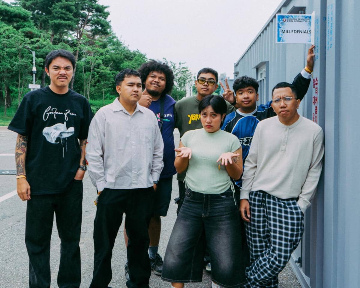 Tujuh anggota grup musik Milledenials berpose santai di area luar ruangan Pentaport Rock Festival 2025 dengan latar pepohonan hijau.